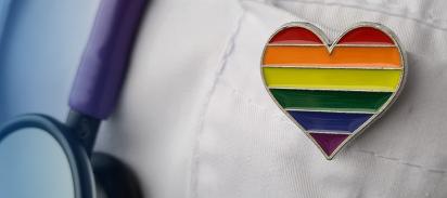 Anstecker in Form eines Herzens in Regenbogenfarben steckt an einem Arztkittel, daneben ein Stetoskop.