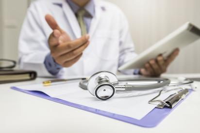Arzt im Gespräch, hält einen Schreibblock in der linken Hand. Im Vordergrund des Bildes liegt ein Stetoskop und Klembrett auf dem Schreibtisch.