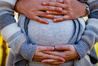 Eine schwangere Person umfasst den Bauch mit ihren Händen, von hinten wir sie von einer anderen Person umfasst.