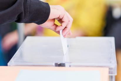 Eine Person, zu sehen ist nur der Unterarm mit der Hand, steckt einen Stimmzettel in eine Wahlurne